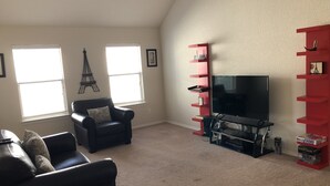 Una sala de estar con un sofá de cuero negro, un mueble de entretenimiento rojo, un televisor de pantalla plana y dos ventanas con persianas.