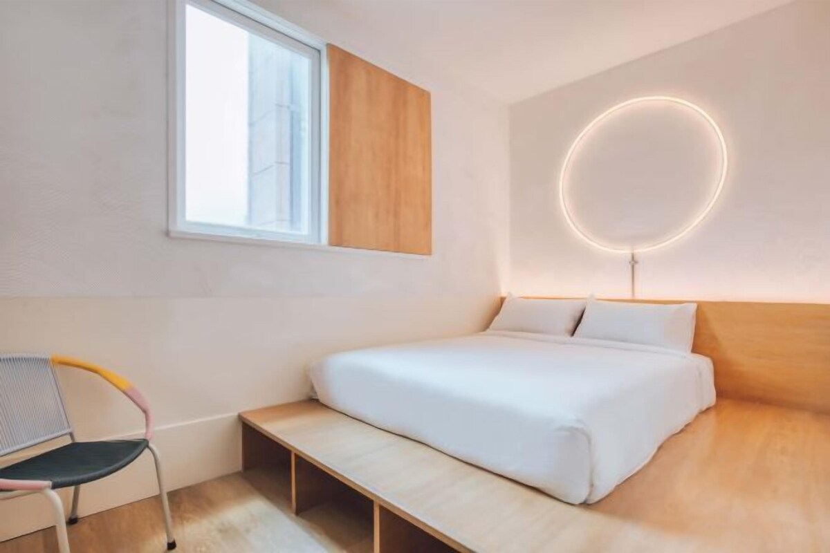 A modern bedroom with a bed, a bench, a chair, a window, and a wall-mounted light fixture.