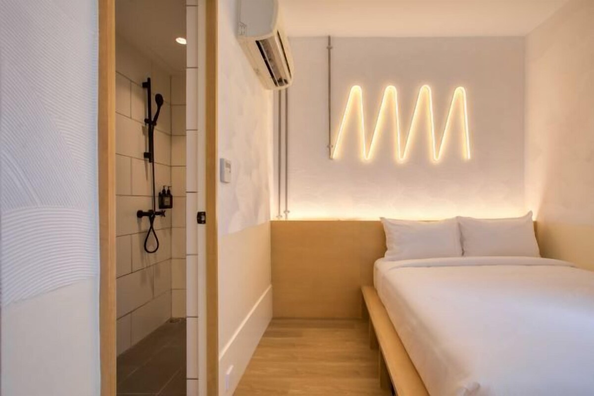 A modern hotel room with a wooden headboard, a bed with white linens, a shower area with a handheld showerhead, and a wall-mounted air conditioning unit.