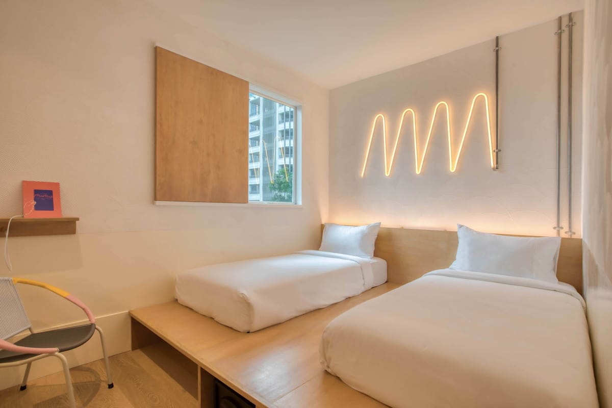 A modern hotel room with two single beds, a wooden bench, a chair, and a large window.