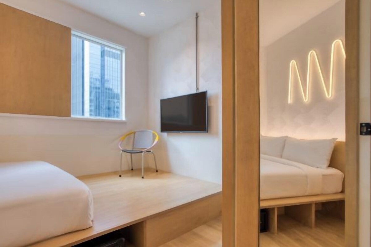 A modern hotel room with a bed, a desk, a chair, a TV, and a neon sign.