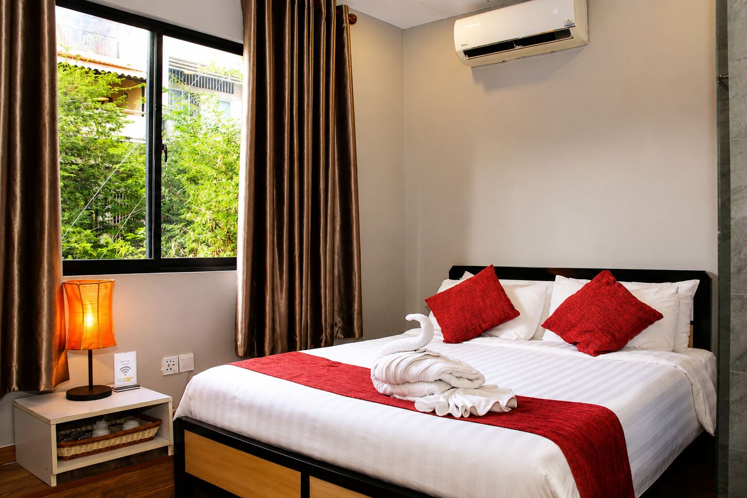 A hotel room with a bed, a nightstand, a lamp, a window with curtains, and an air conditioning unit.