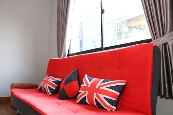 A red sofa with British flag cushions, a window with curtains, and a view of buildings outside. - Ostro Hotel (Phnom Penh)