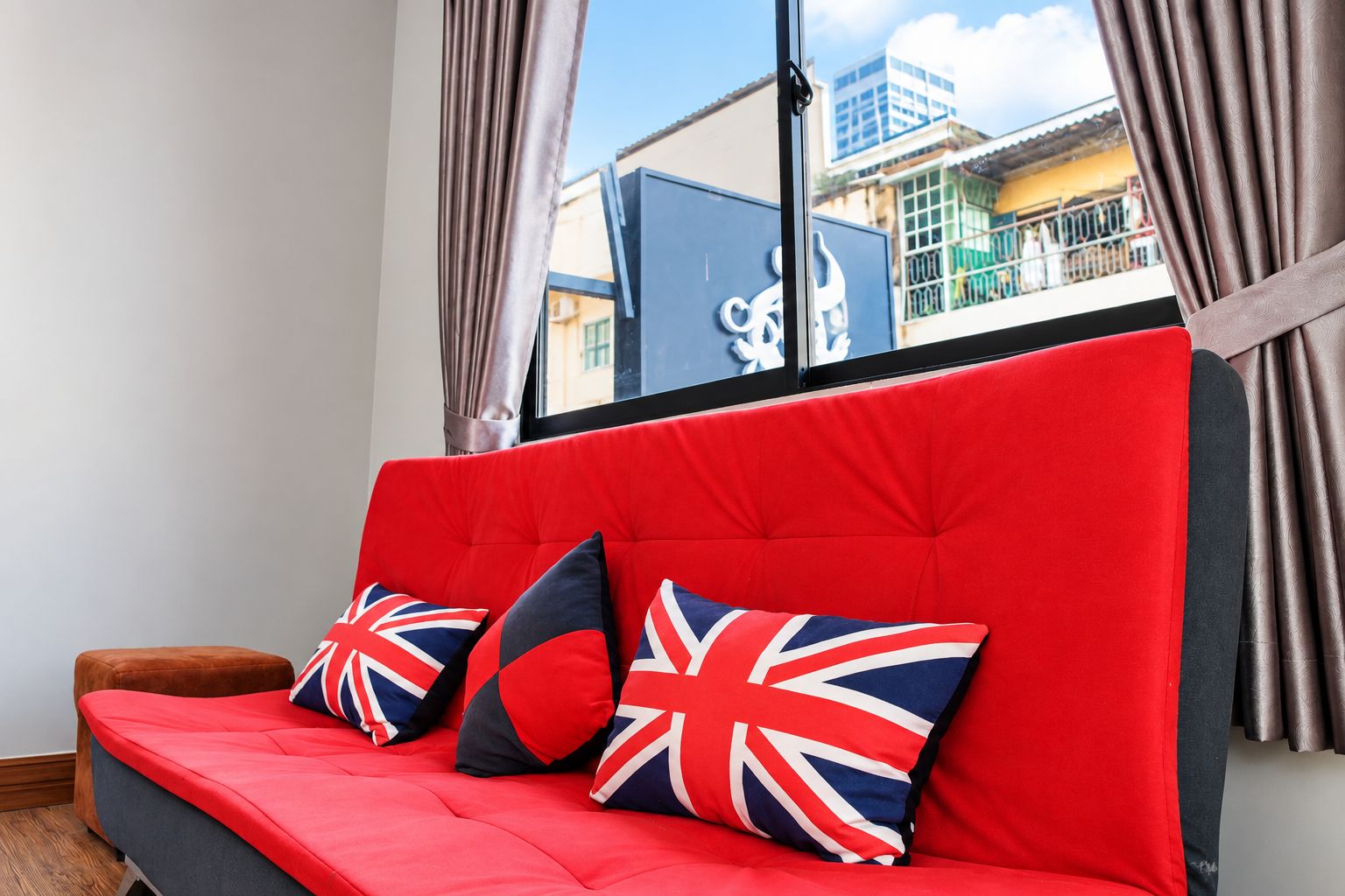 A red sofa with British flag cushions in a room with a view of buildings through a window.