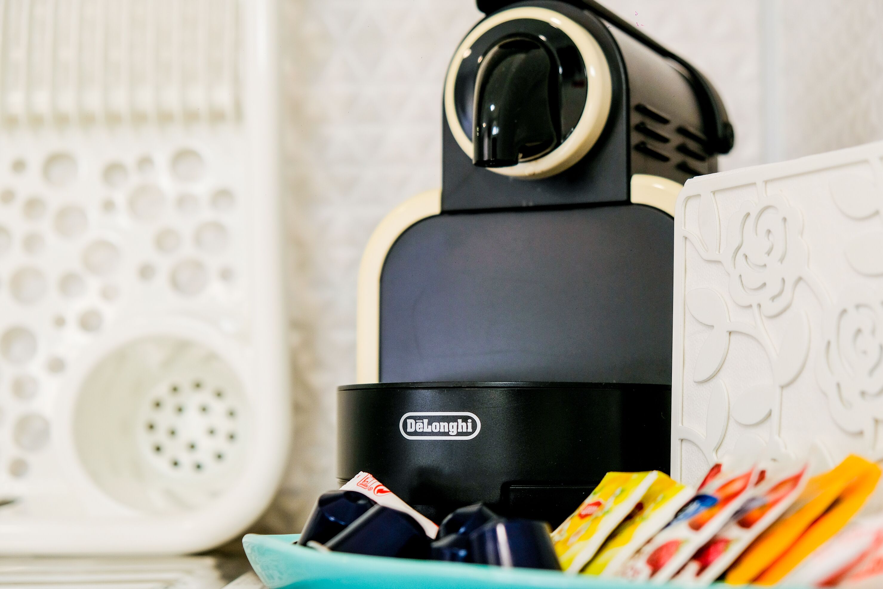 Eine DeLonghi-Espressomaschine mit türkisem Sockel, eine weiße Keramiktasse mit bunten Gegenständen und ein weißes Keramikobjekt mit geprägten Mustern.