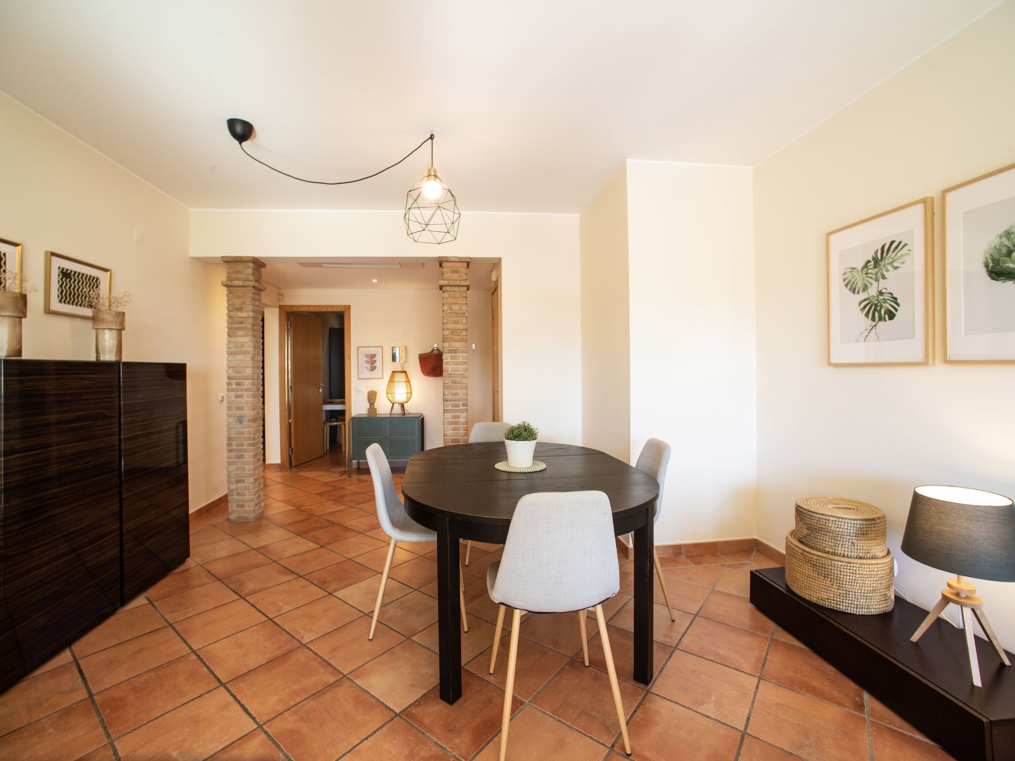 A dining area with a round table, four chairs, a hanging light fixture, and a brick wall.