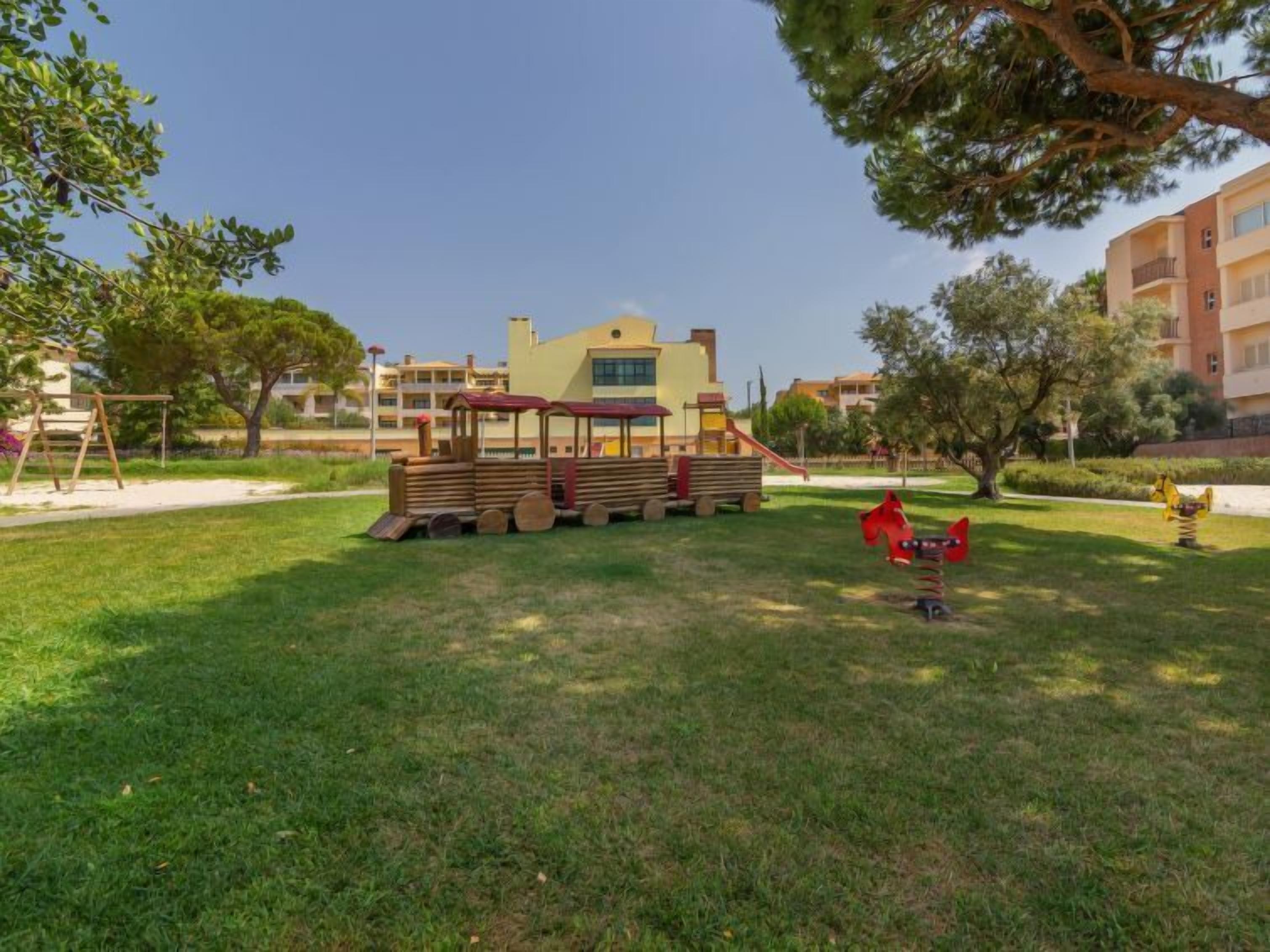 Un parque infantil con un paseo en tren y columpios en un área verde, con edificios al fondo.