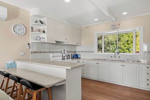 A kitchen with white cabinets, a central island, bar stools, a clock, and a window with a view of greenery.