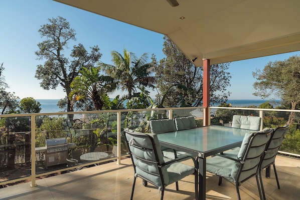 A glass-top table with four chairs, a grill, and a view of the sea.
