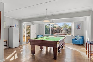 A spacious room with a pool table, a refrigerator, a blue armchair, and a large window offering a view of the outdoors.