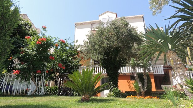 A building with a white facade, a balcony, and a garden with various plants and flowers. - Apartments Travarevic (Dubrovnik)