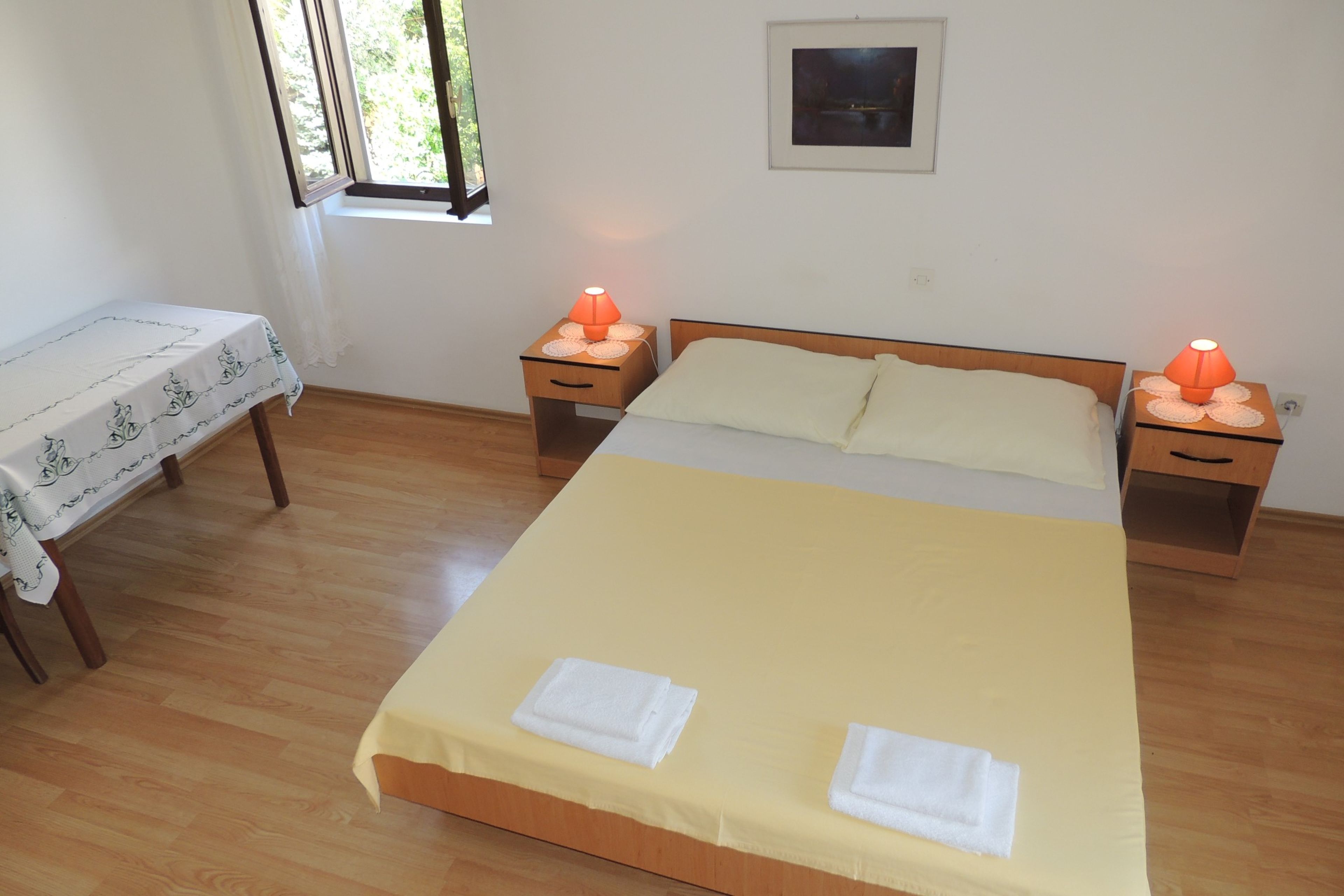 Un dormitorio con cama, dos mesitas de noche con lámparas, una ventana, una pequeña mesa con mantel y un cuadro en la pared.