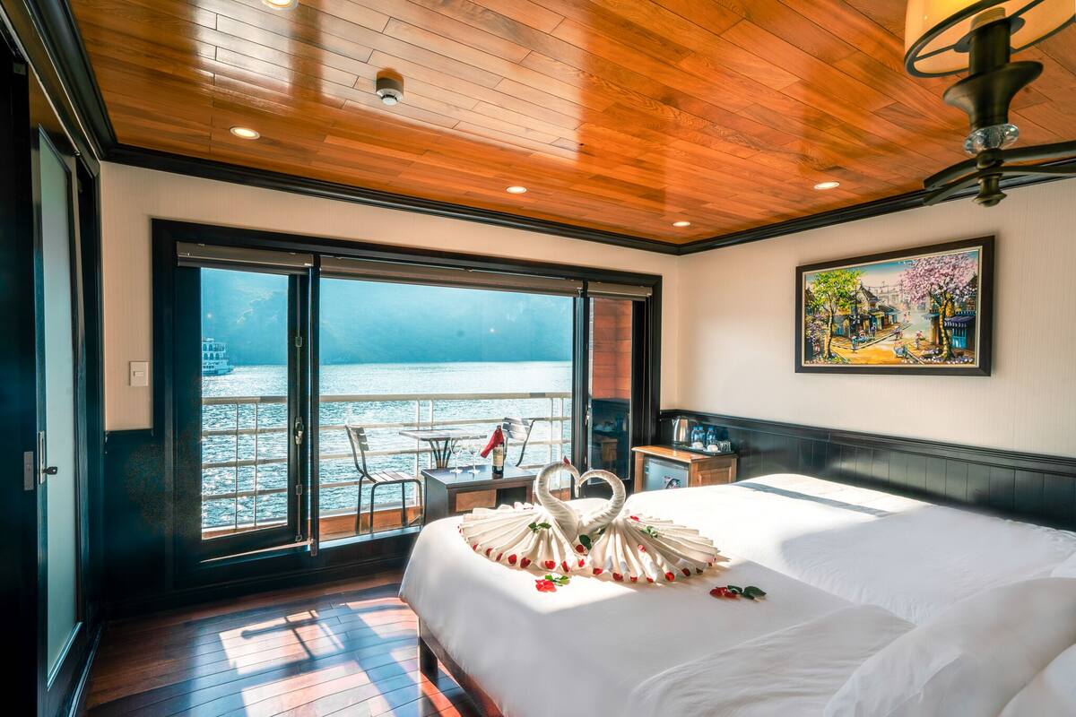 A hotel room with a large bed, a wooden ceiling, a painting on the wall, and a view of the sea through a sliding door.