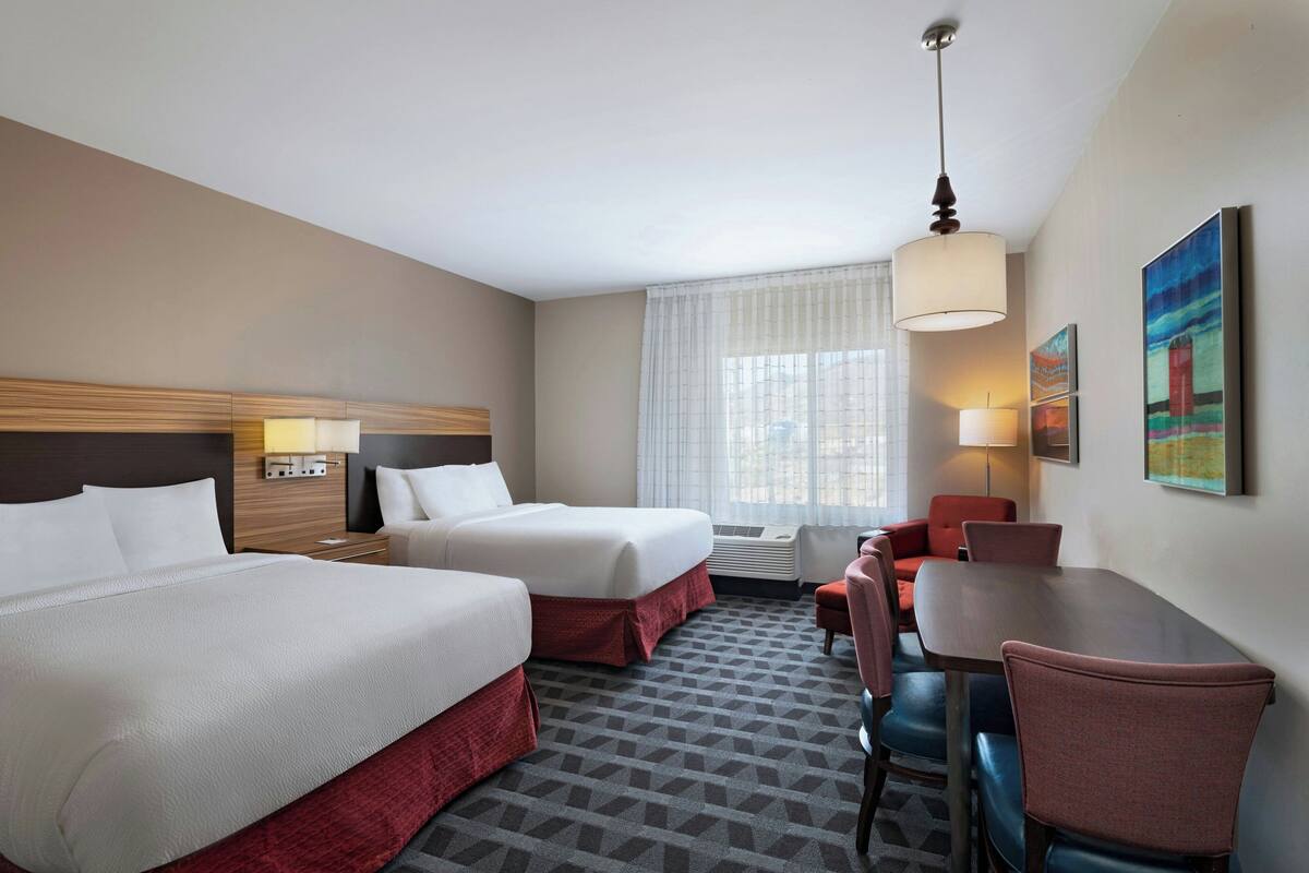 Une chambre d’hôtel avec deux lits, un bureau, des chaises, une lampe et des œuvres d’art accrochées au mur.