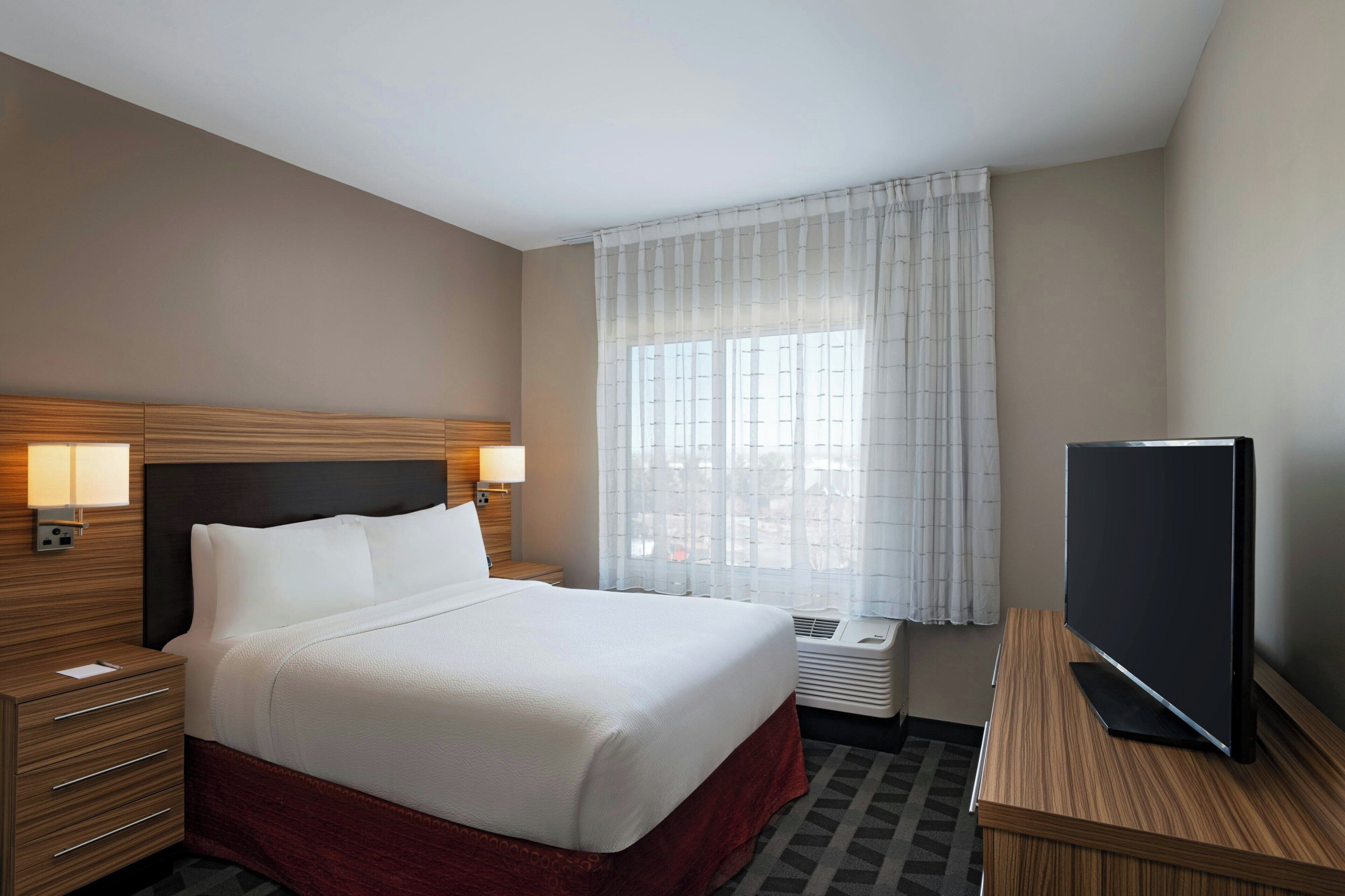 A hotel room with a large bed, bedside lamps, a flat-screen TV, and a window with sheer curtains.