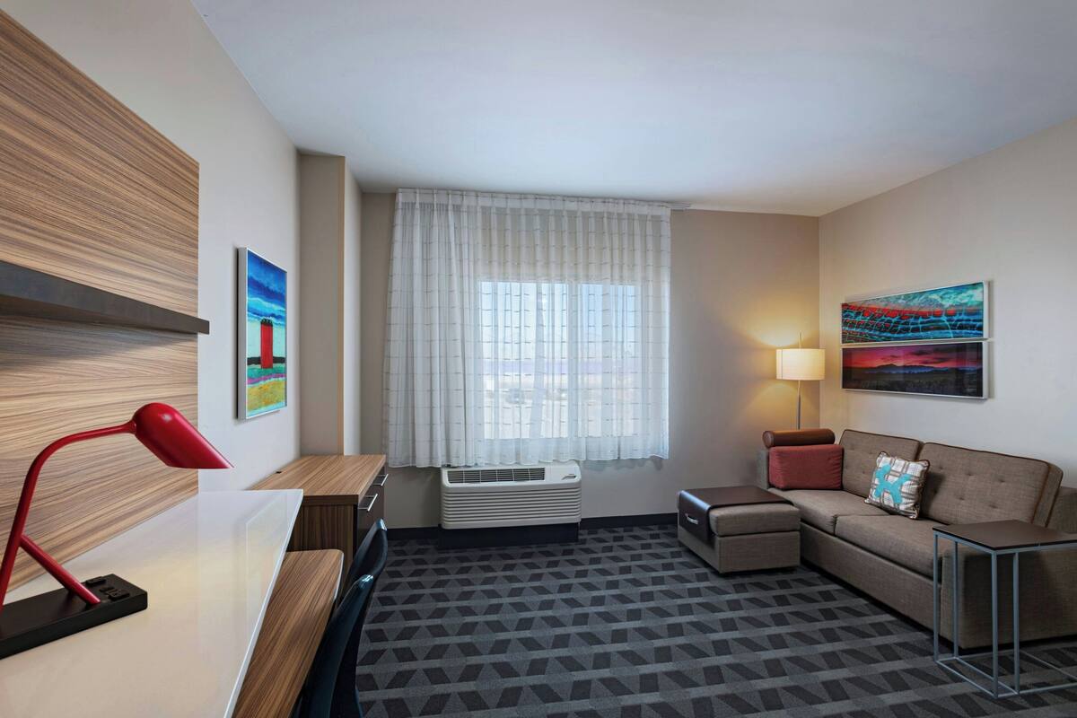 Une chambre d’hôtel avec un bureau, une lampe rouge, un canapé, une petite table, un lampadaire et une grande fenêtre avec des rideaux légers.