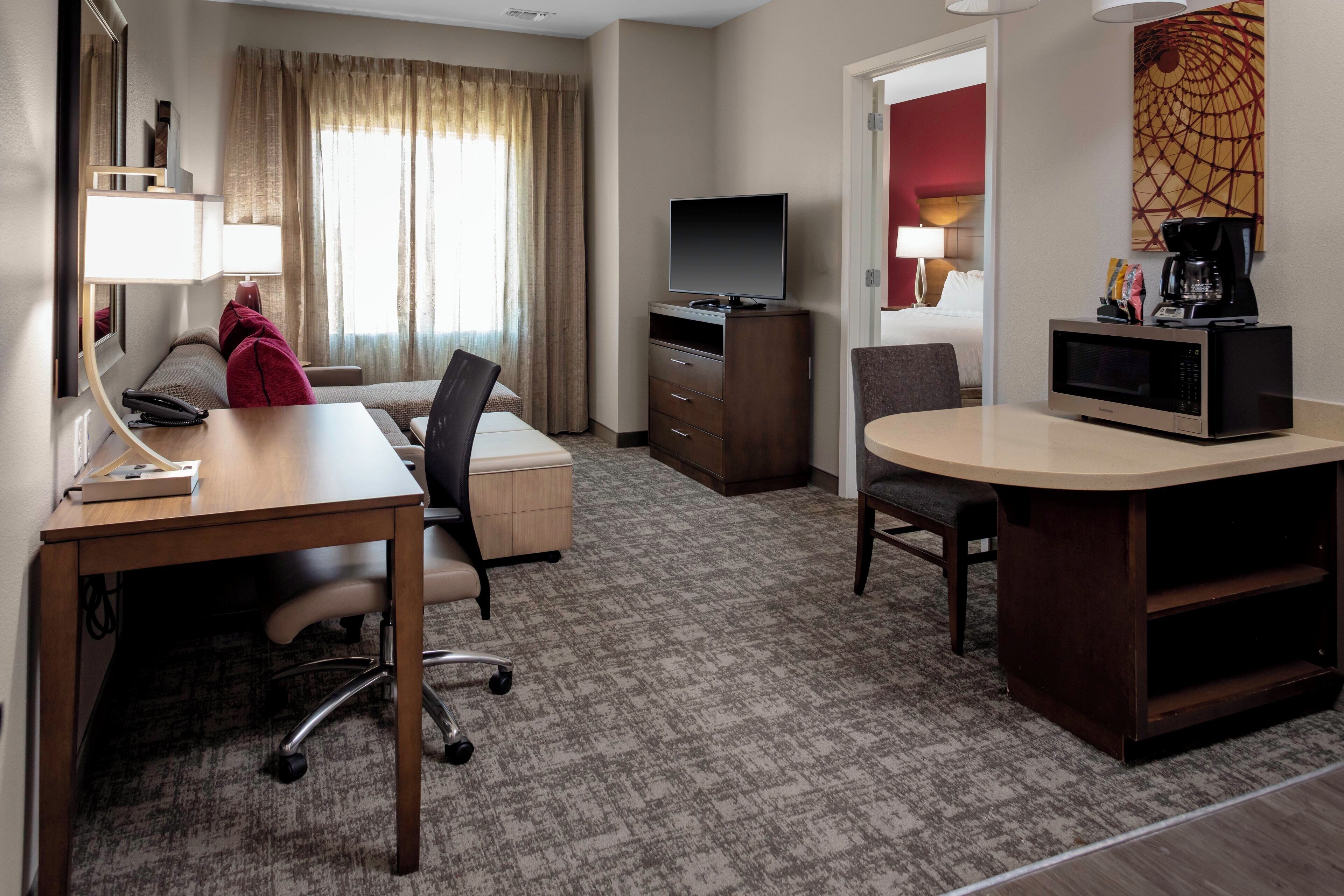 Habitación de hotel con escritorio, silla, televisor, microondas y una cama con cabecera roja.