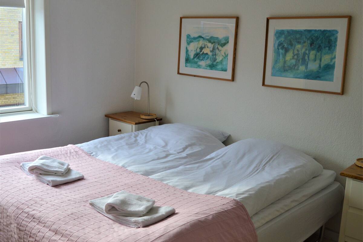 A bedroom with two beds, a window, a nightstand with a lamp, and two framed artworks on the wall.