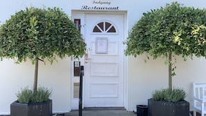 A white door with the sign "Indgang Restaurant" above it. There are two potted trees on either side of the door. - Strandby Badehotel (Strandby)