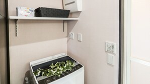 A laundry area with a front-loading washing machine, a shelf with a box and a basket, and a power outlet. - Vacation Rent Kanayama (Nagoya)