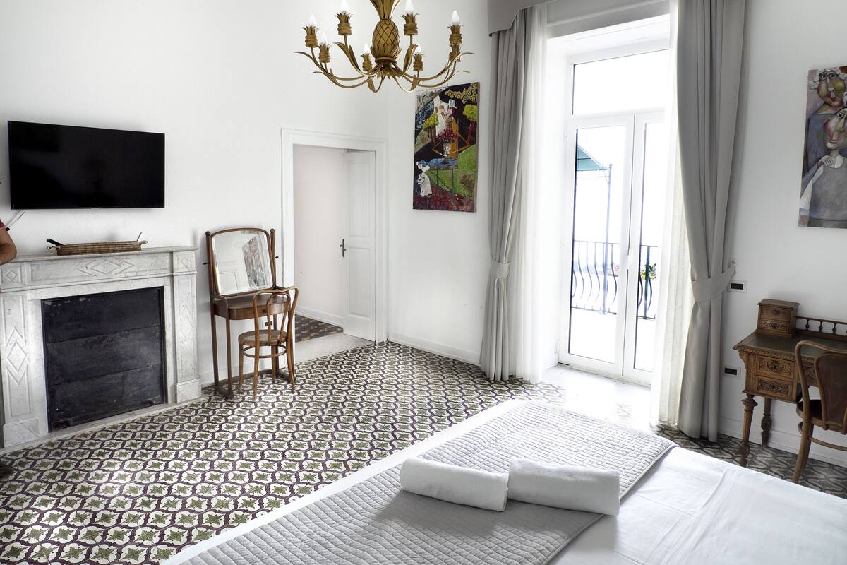 A hotel room with a fireplace, a bed with pillows, a chair, a desk, a television, and a painting on the wall.