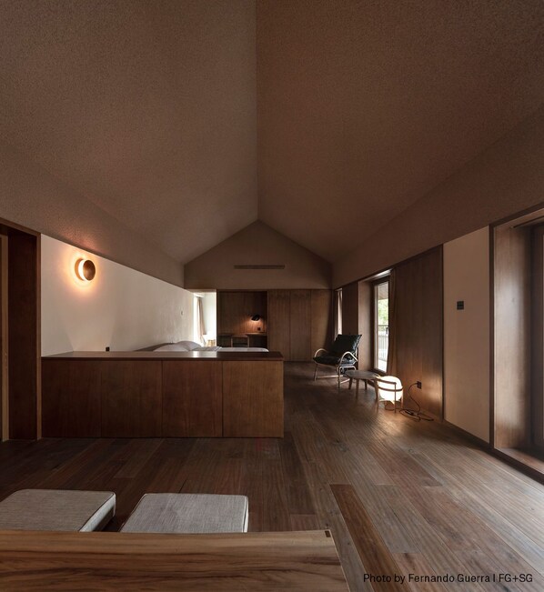 A spacious, modern hotel room with a wooden floor, a central island bed, a sitting area with a chair, and large windows providing natural light.