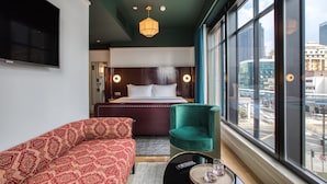 Een hotelkamer met een groot bed, een rode bank met patroon, een groene fauteuil, een aan de muur bevestigde televisie en uitzicht op de stad door grote ramen.