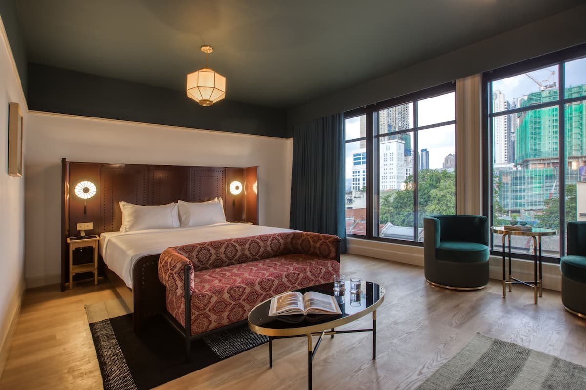 Une chambre d’hôtel moderne, dotée d’un grand lit, d’un canapé, d’une petite table et offrant une vue sur la ville grâce à de grandes fenêtres.