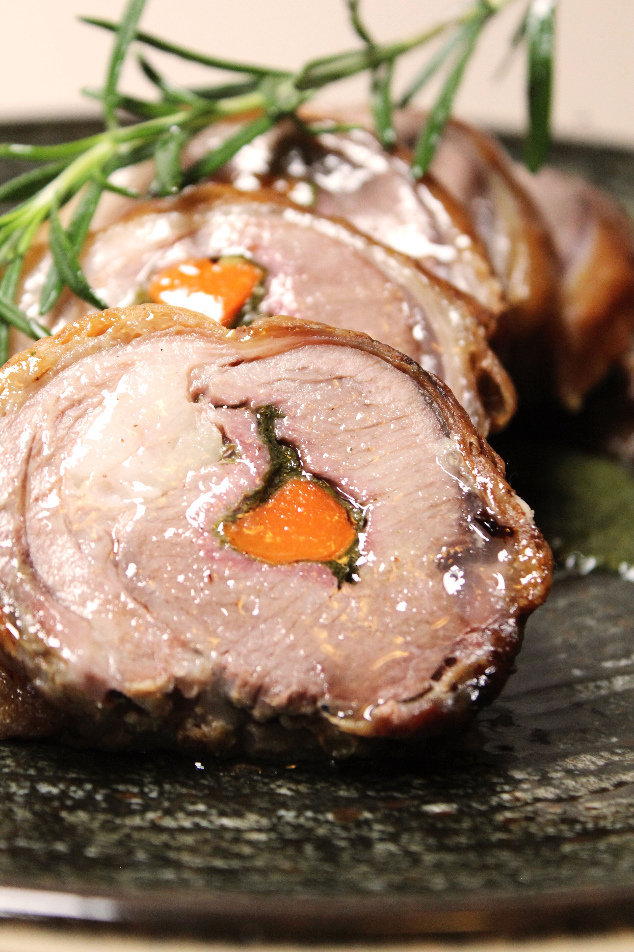 A close-up of sliced meat with herbs and a carrot, served on a dark plate.