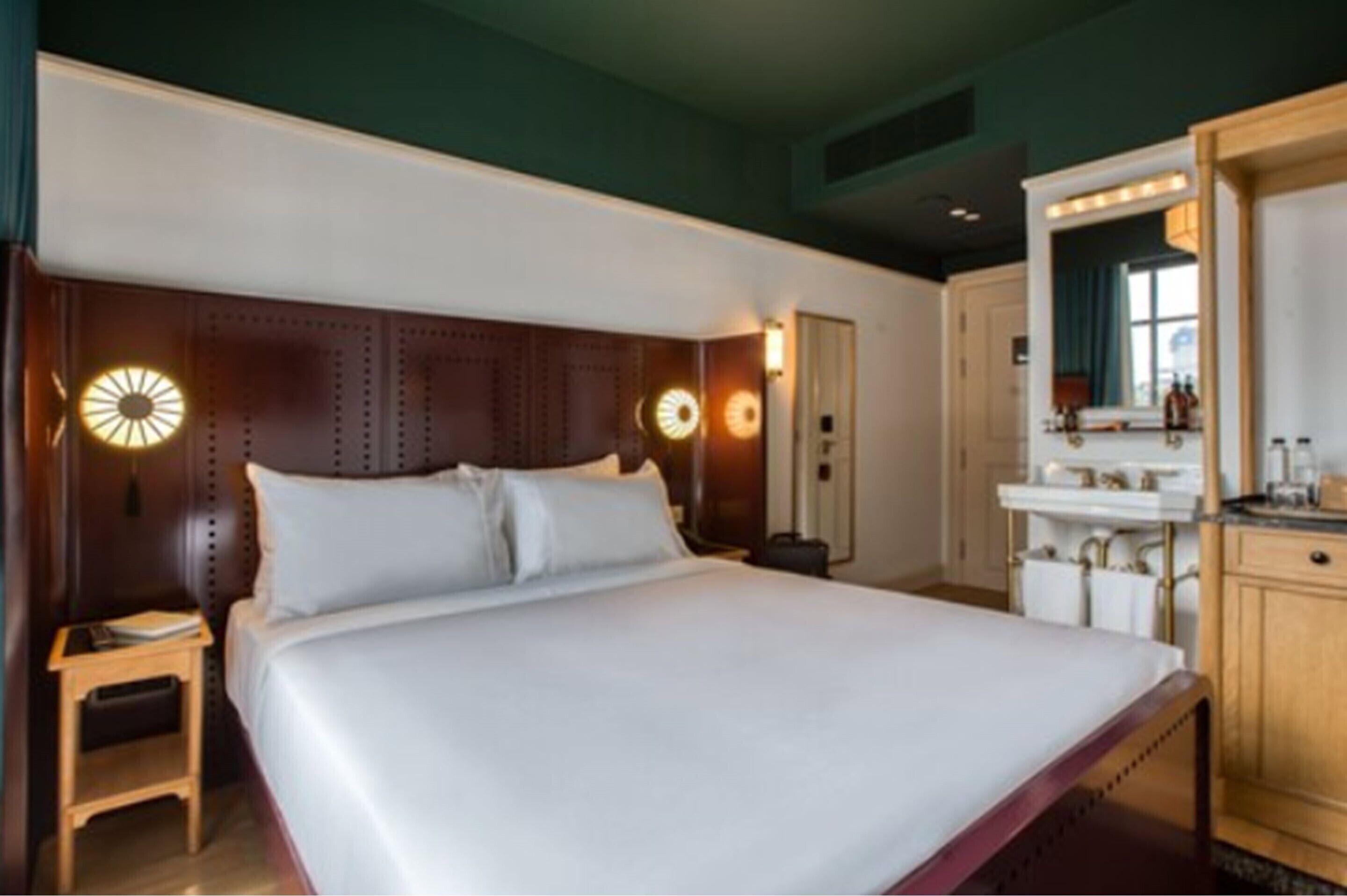 A hotel room with a large bed, a wooden headboard, a vanity with a mirror, and a small table with a lamp.