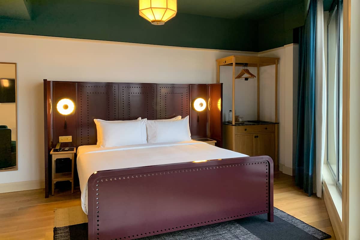 Une chambre d’hôtel moderne avec un grand lit, une tête de lit en bois, des lampes de chevet, une petite table de chevet, un miroir et une fenêtre avec des rideaux.