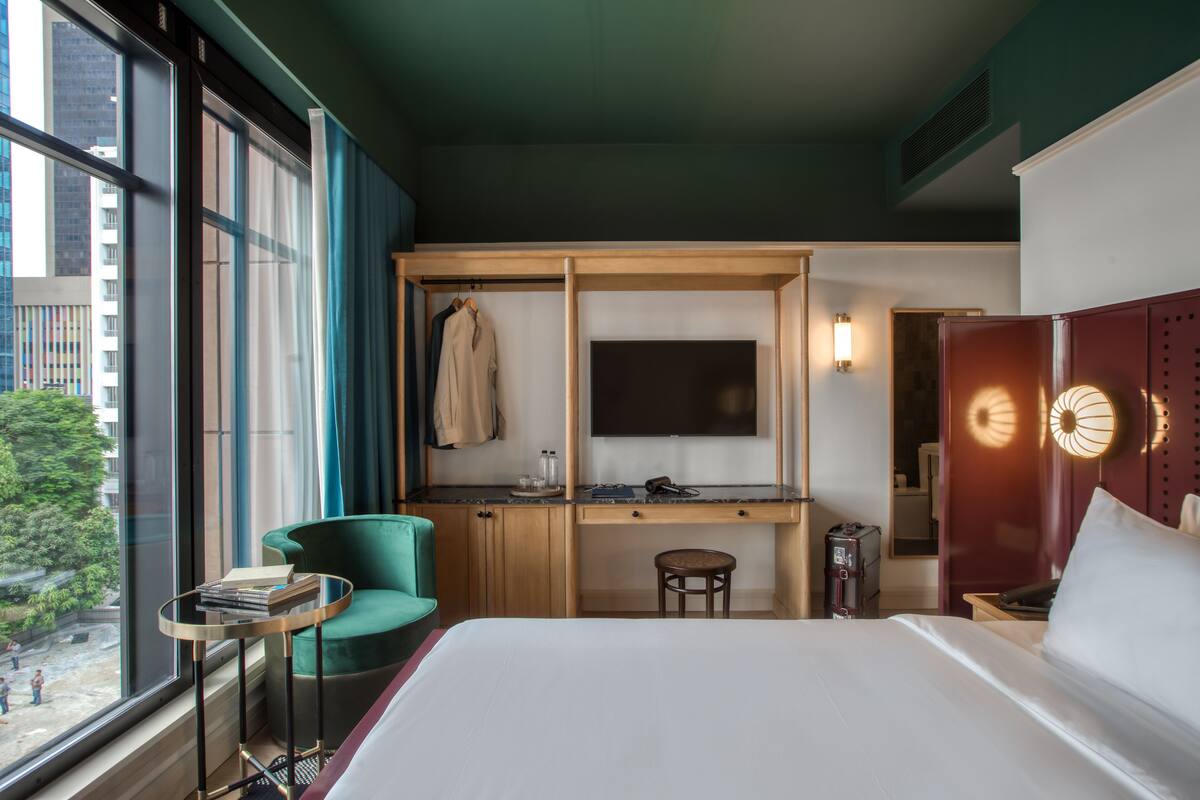 Une chambre d’hôtel équipée d’un lit, d’un bureau, d’une chaise, d’une télévision et d’une fenêtre donnant sur des immeubles et des arbres.
