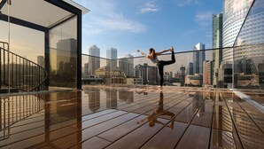 Een persoon doet een yoga-houding op een houten terras, met een stadsgezicht op de achtergrond.