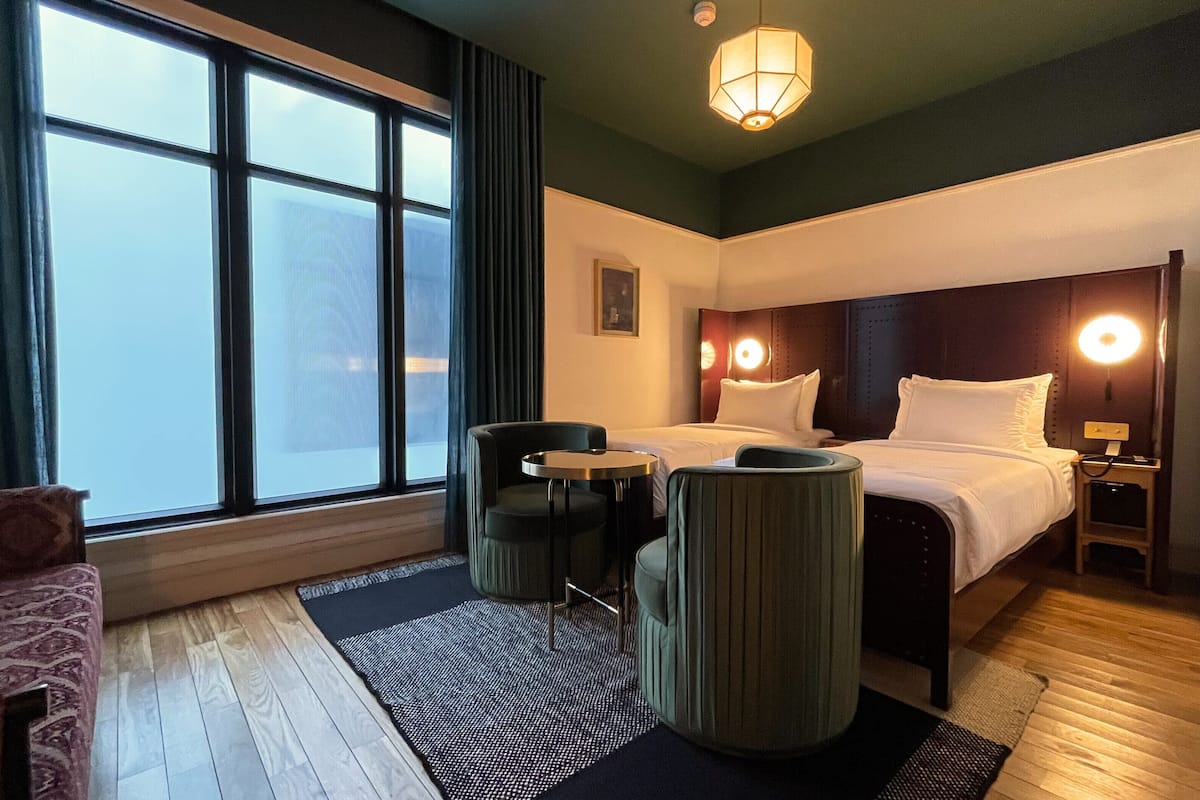 Une chambre d’hôtel avec une grande fenêtre, un lit, deux chaises, une petite table et une table de chevet avec une lampe.