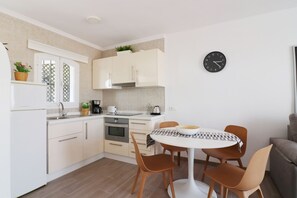 A modern kitchen with white cabinets, a dining table with four chairs, a clock on the wall, and a grey sofa in the corner.