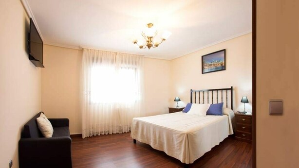A bedroom with a bed, a nightstand, a lamp, a chair, a window with curtains, and a chandelier.