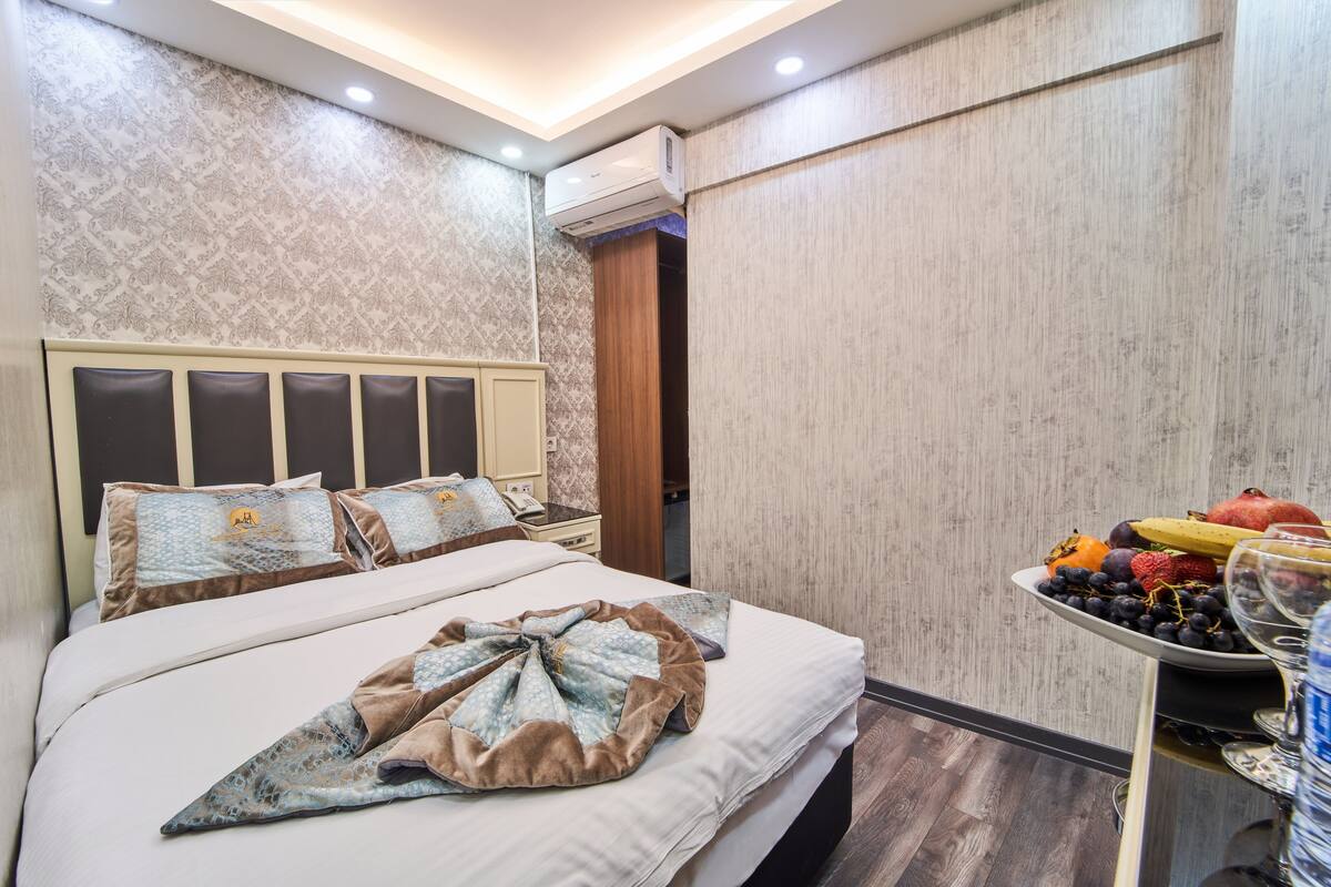 A hotel room with a large bed, a bedside table with fruit and glasses, and a wall-mounted air conditioner.