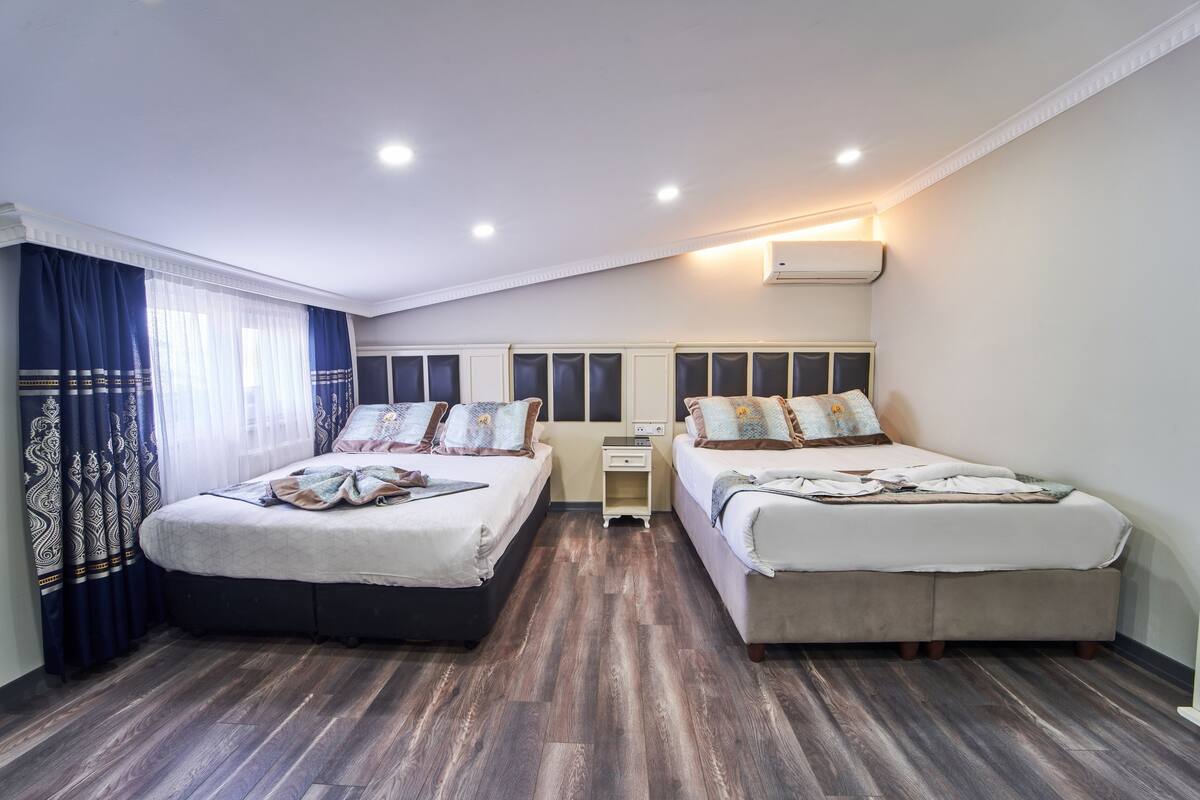 A hotel room with two beds, a small bedside table, a nightstand, and a ceiling-mounted air conditioner.