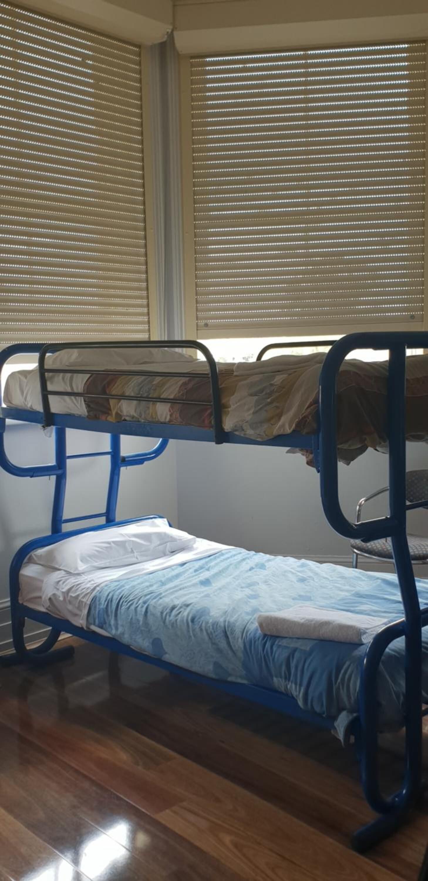 A bunk bed with a blue metal frame, a white mattress, and a blue blanket. There are two windows with blinds, and the room has wooden flooring.