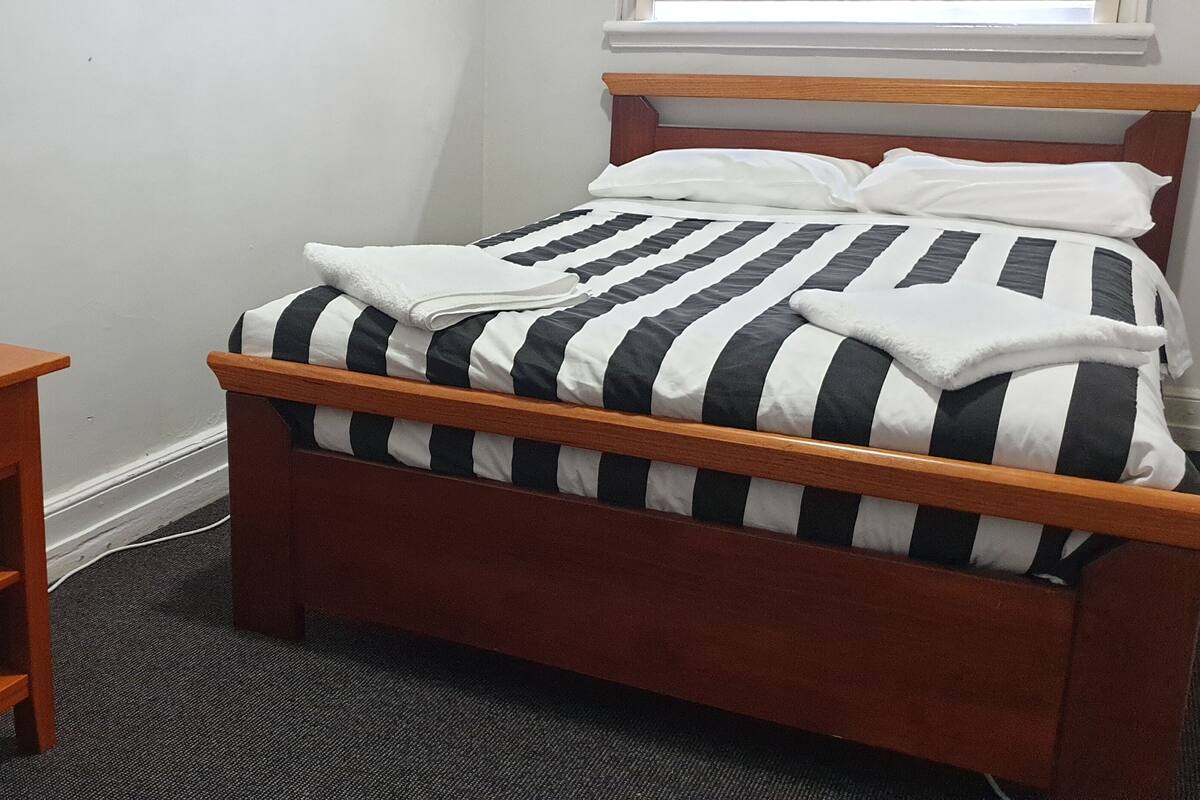 A neatly made bed with striped bedding, a bedside table with a lamp, and a small drawer unit.
