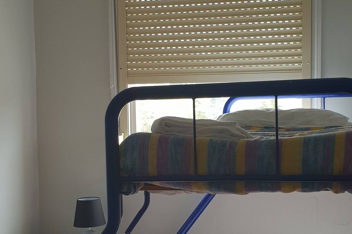 A bunk bed with a striped blanket, a bedside table with a lamp, and a window with blinds.