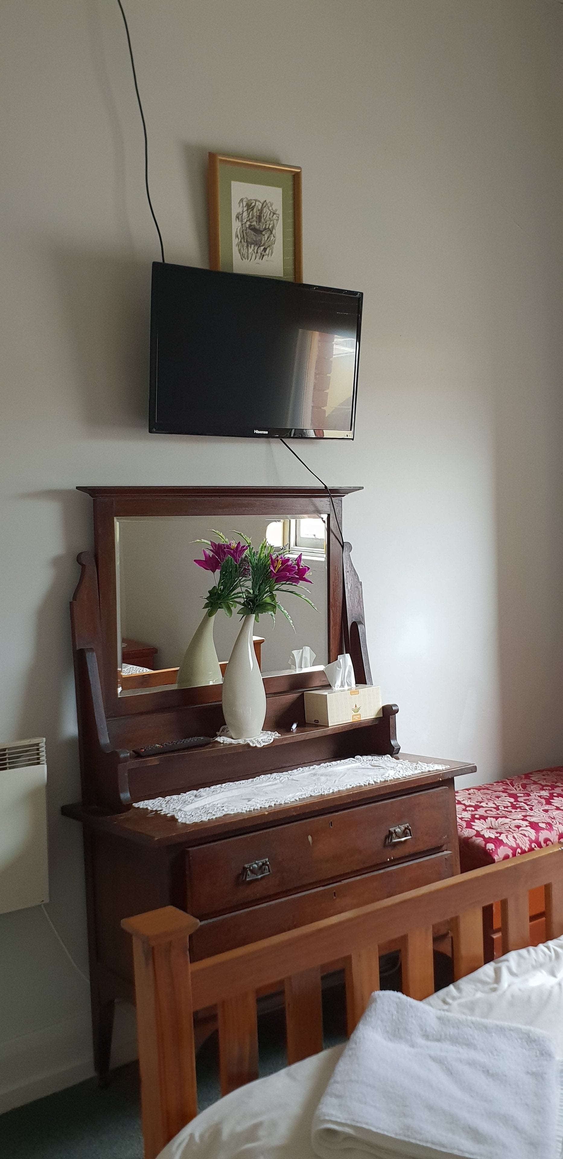 Une commode en bois avec un miroir, un vase de fleurs et un téléviseur fixé au mur au-dessus.