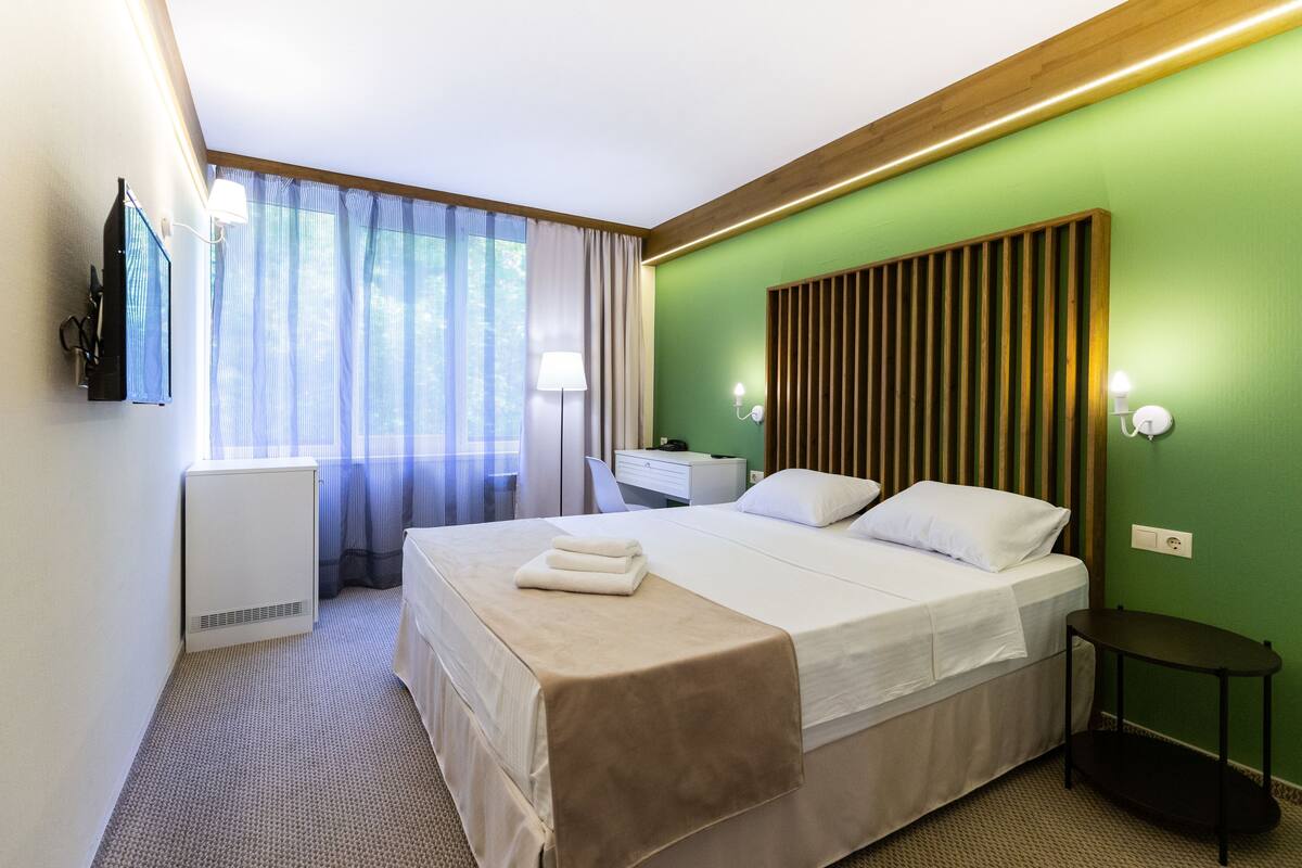 A hotel room with a bed, a desk, a chair, a lamp, and a TV.