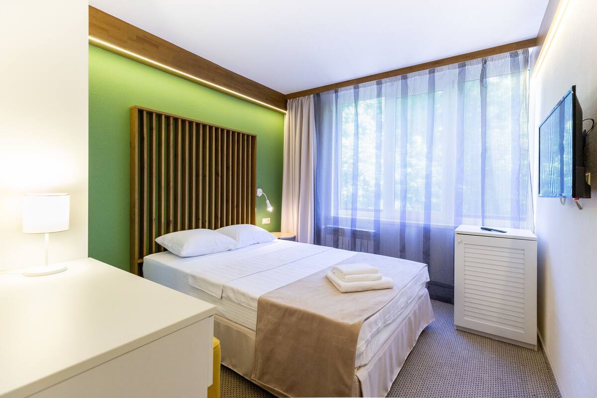 A hotel room with a bed, a desk, a lamp, a TV, and a window with sheer curtains.