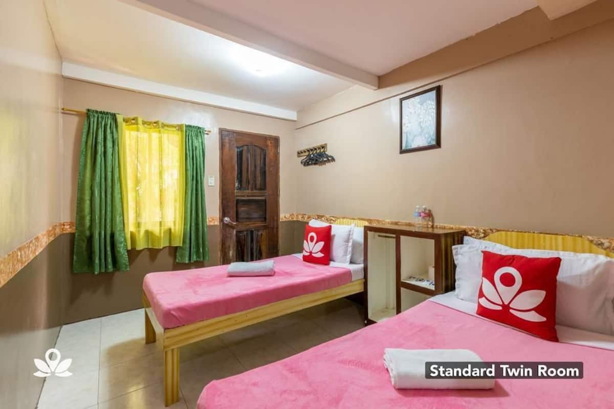 Ein Standard-Doppelzimmer mit zwei Betten, einer Holztür, einem kleinen Tisch und einem Fenster mit grün-gelben Vorhängen.