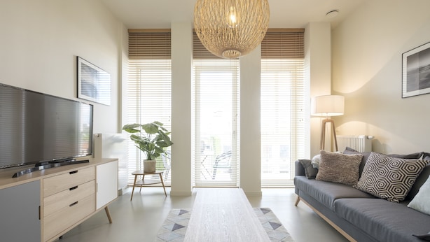 Una sala de estar moderna con un televisor de pantalla plana, un sofá gris, una mesa de centro de madera y un ventanal con persianas.