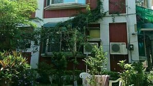 Ein zweistöckiges Gebäude mit roter und weißer Fassade, ein kleiner Garten mit Tisch und Stühlen und ein Baum, der Schatten spendet.