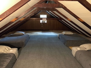 Une chambre de type dortoir avec quatre lits disposés en rangée, un plafond en bois et un luminaire apparent.