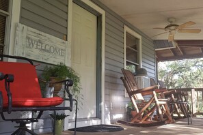 Une véranda avec une chaise rouge, des fauteuils à bascule en bois, une pancarte de bienvenue et des plantes en pot.