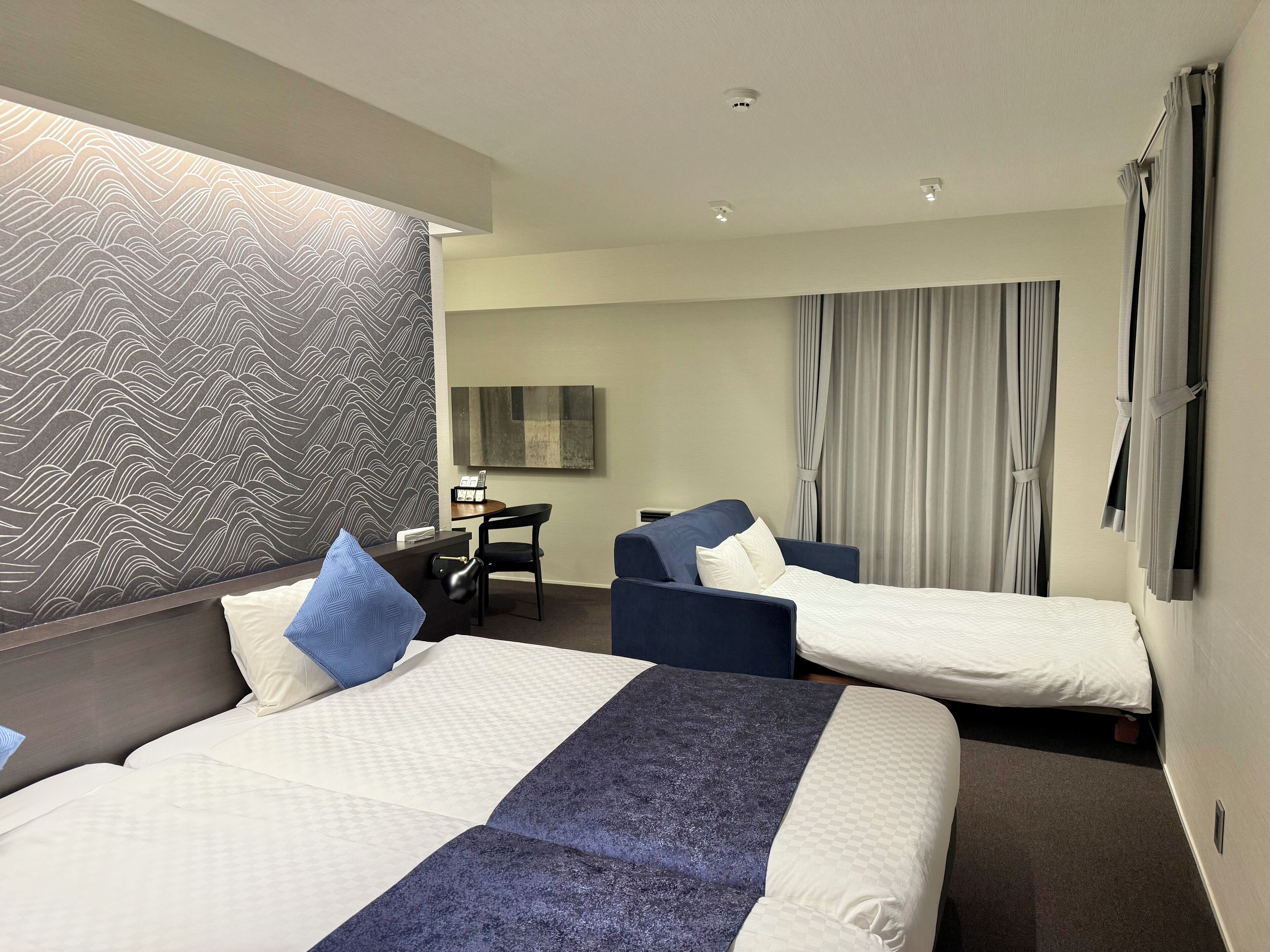 A hotel room with two beds, a desk, a chair, a large mirror, and a view of the outside through curtains.